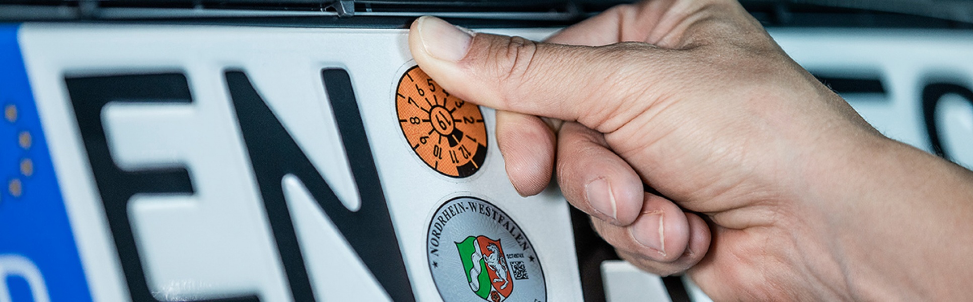 KFZ-Kennzeichen auf die eine Hand eine frische TÜV-Plakette klebt